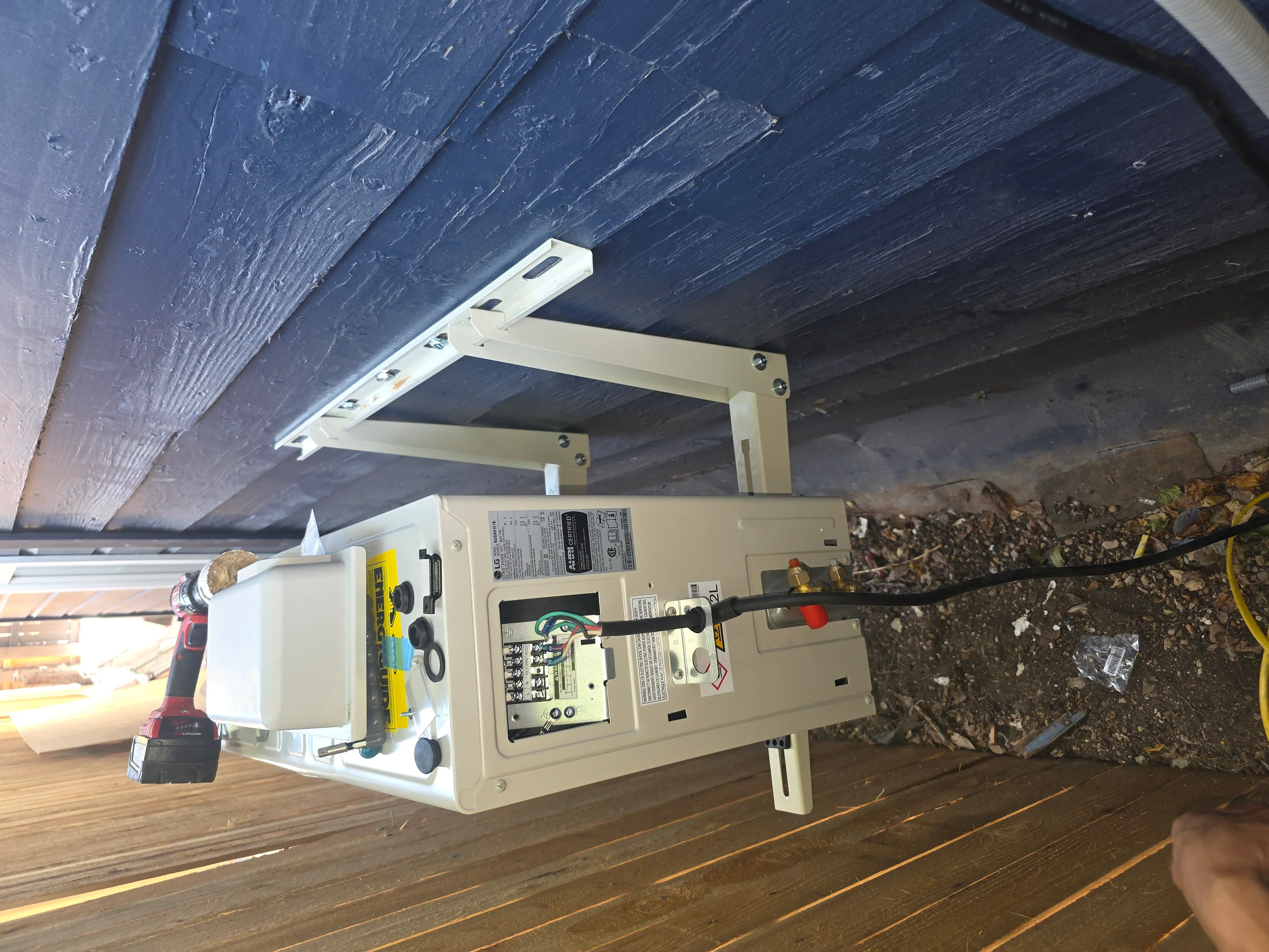 Electrical subpanel mounted to floor joists with white brackets, breakers, and conduit connections